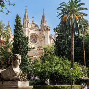 View of the Catedral de Mallorca ('La Seu') framed by palm trees and a stone sphinx statue in Palma on the island of Mallorca in Spain.