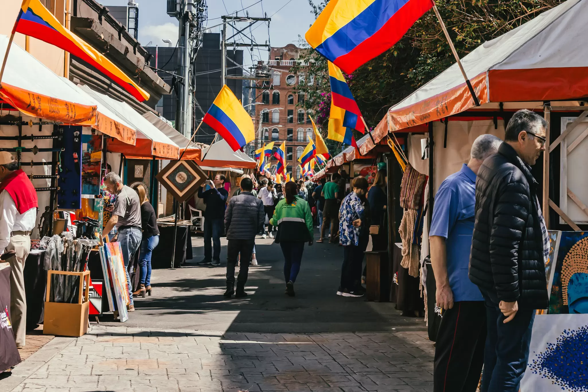 People walk past stalls at an outdoor market; flags with red, yellow and blue stripes hang from each stall.