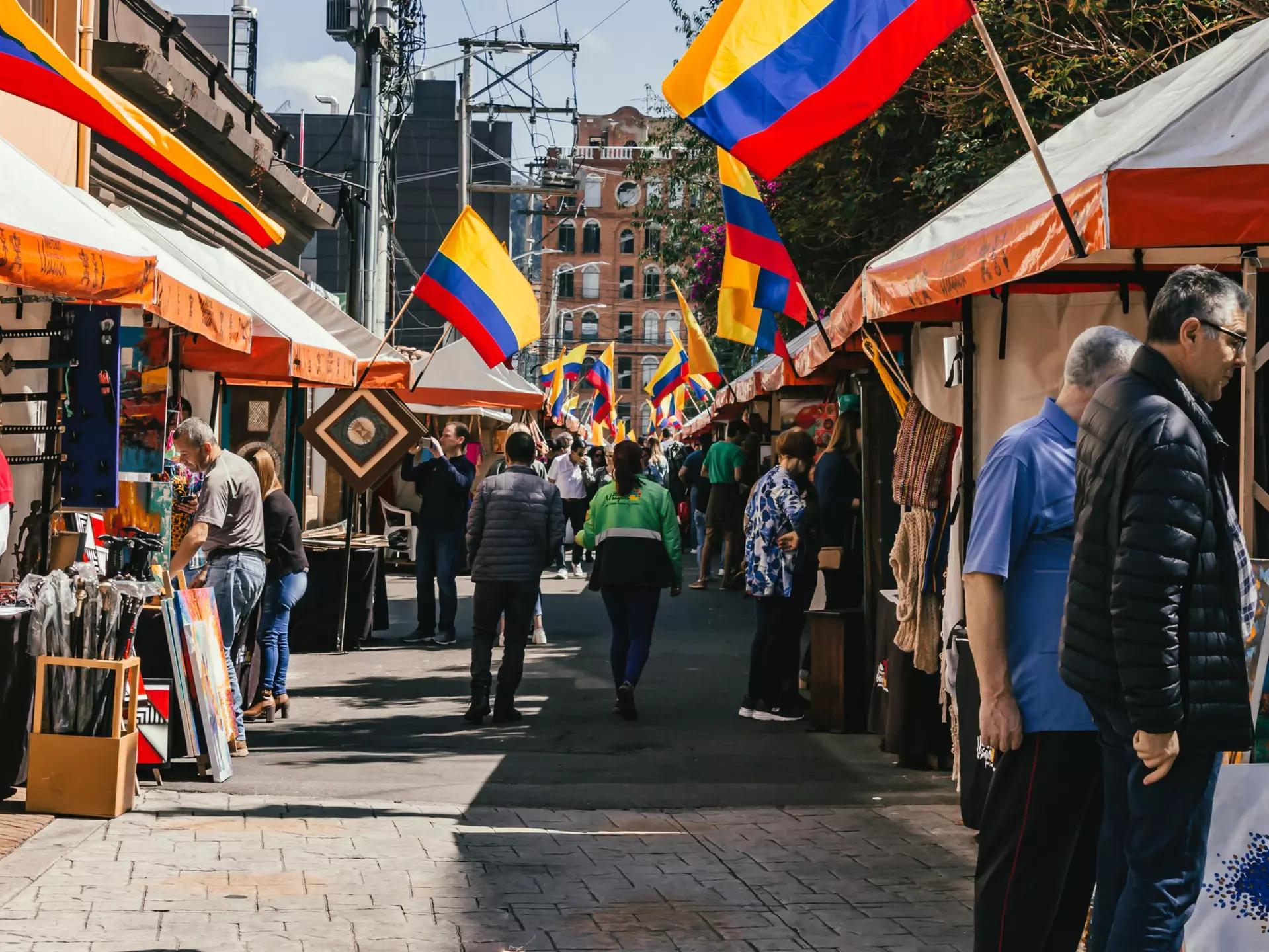 People walk past stalls at an outdoor market; flags with red, yellow and blue stripes hang from each stall.