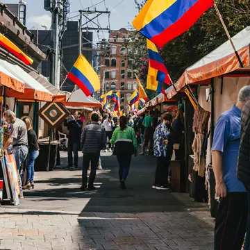People walk past stalls at an outdoor market; flags with red, yellow and blue stripes hang from each stall.