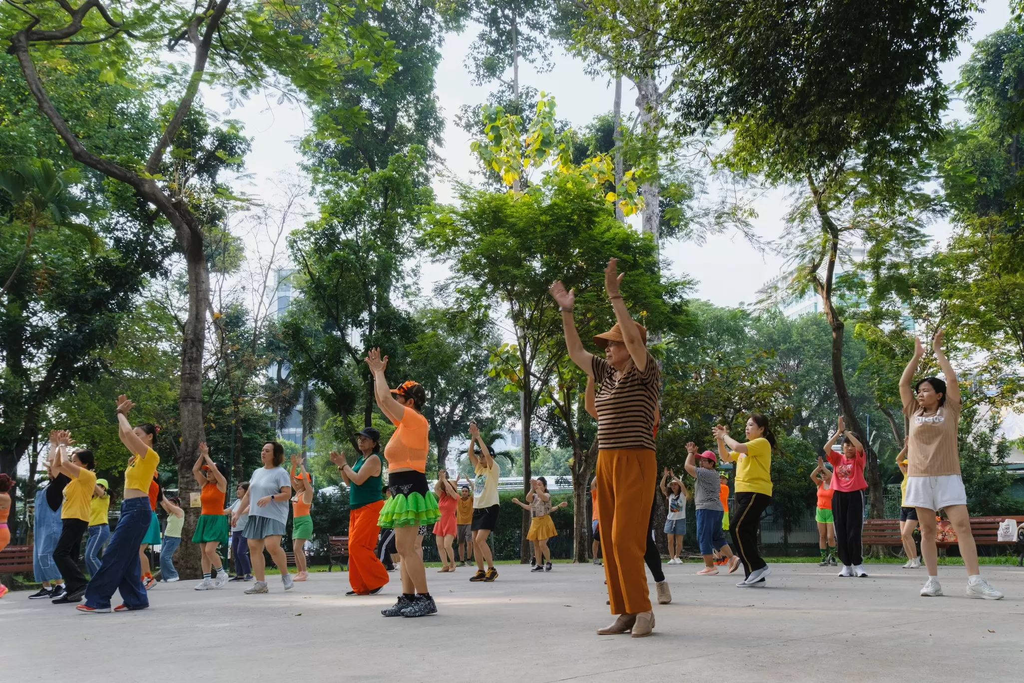 A group of people in brightly colored clothing stand with their hands up in a park, seemingly dancing or stretching