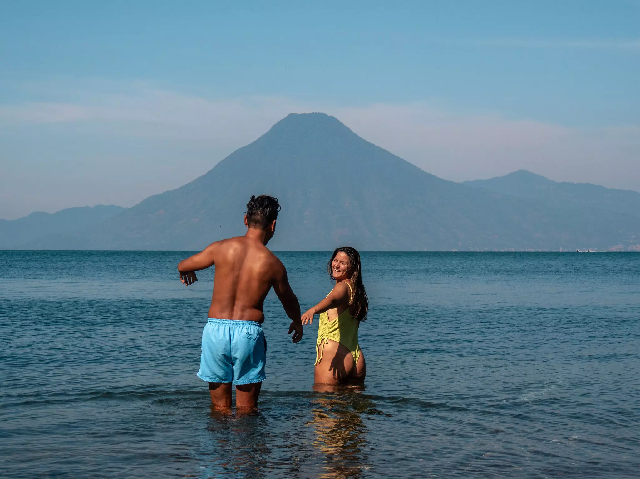 A man and woman in bathing suits wade into the water of Lake Atitlán with a volcano in the distance, Guatemala