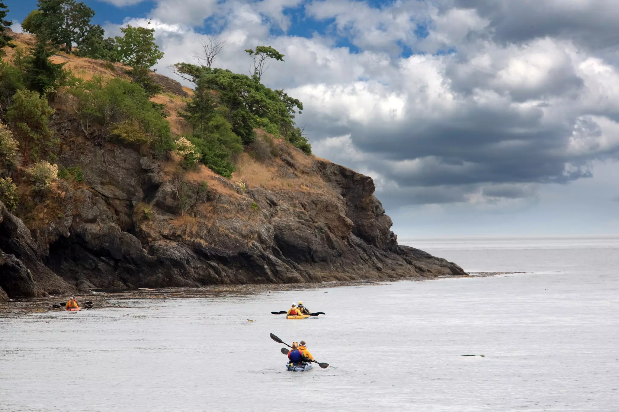 Kayakers in pairs paddle through the ocean near a rocky coastline.