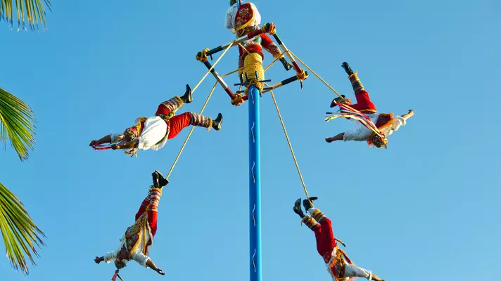 Five acrobat performers attached by rope to a tall blue pole.