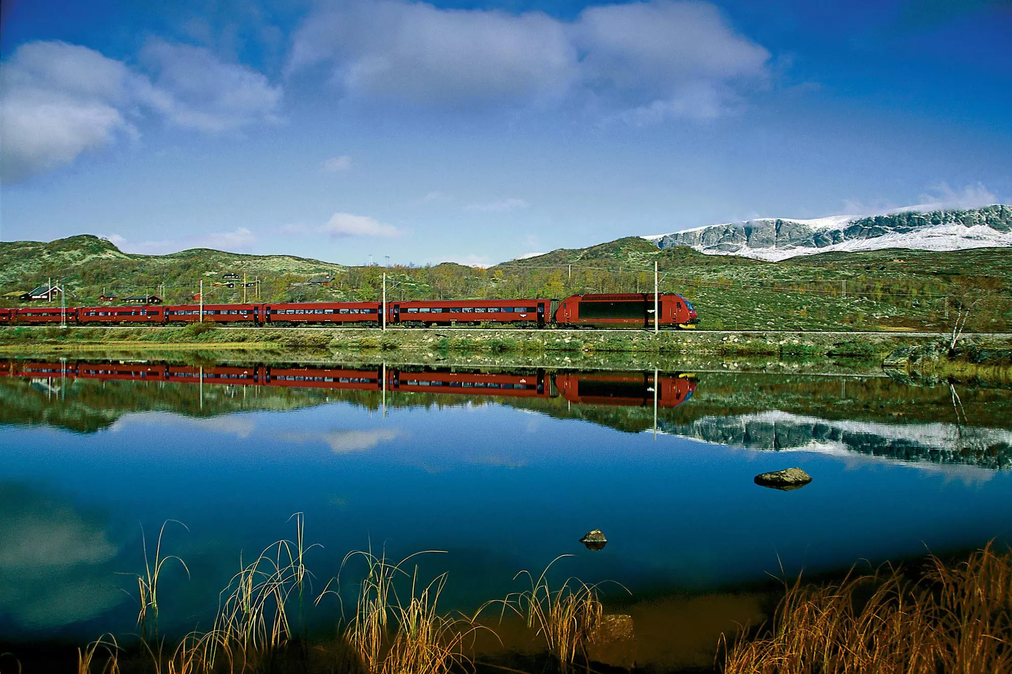 A red train is reflected in the calm surface of a blue lake.