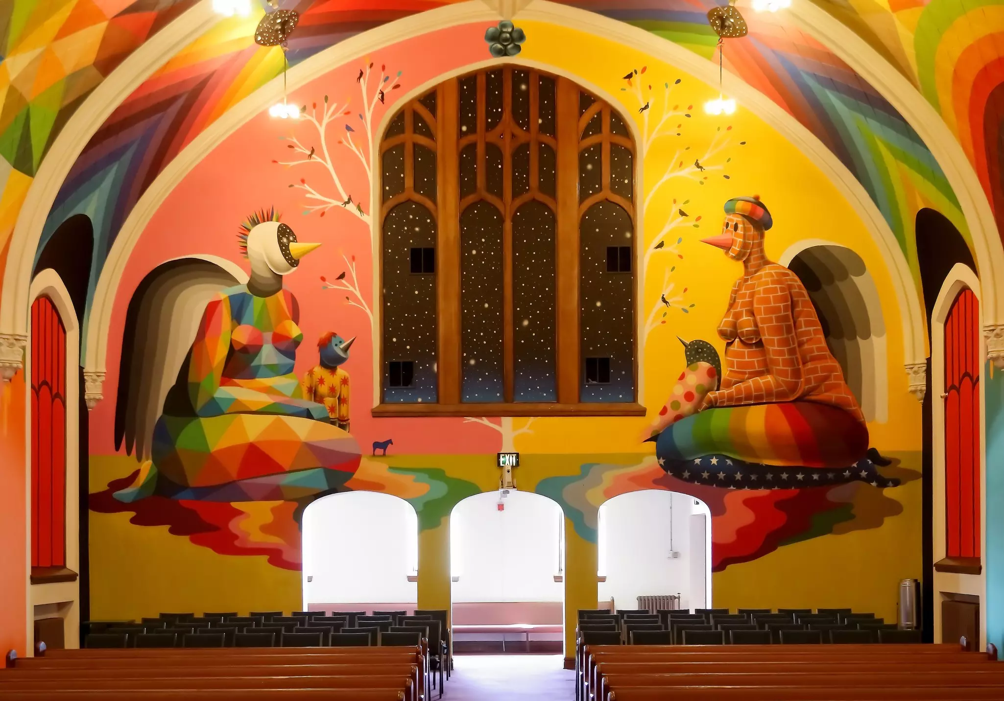 A man with glasses smokes a joint while gazing slightly down at the viewer. He is framed by the rainbow arched ceiling of the International Churhc of Cannabis. Behind him are other people sitting in pews for an Elevationist non denominational service