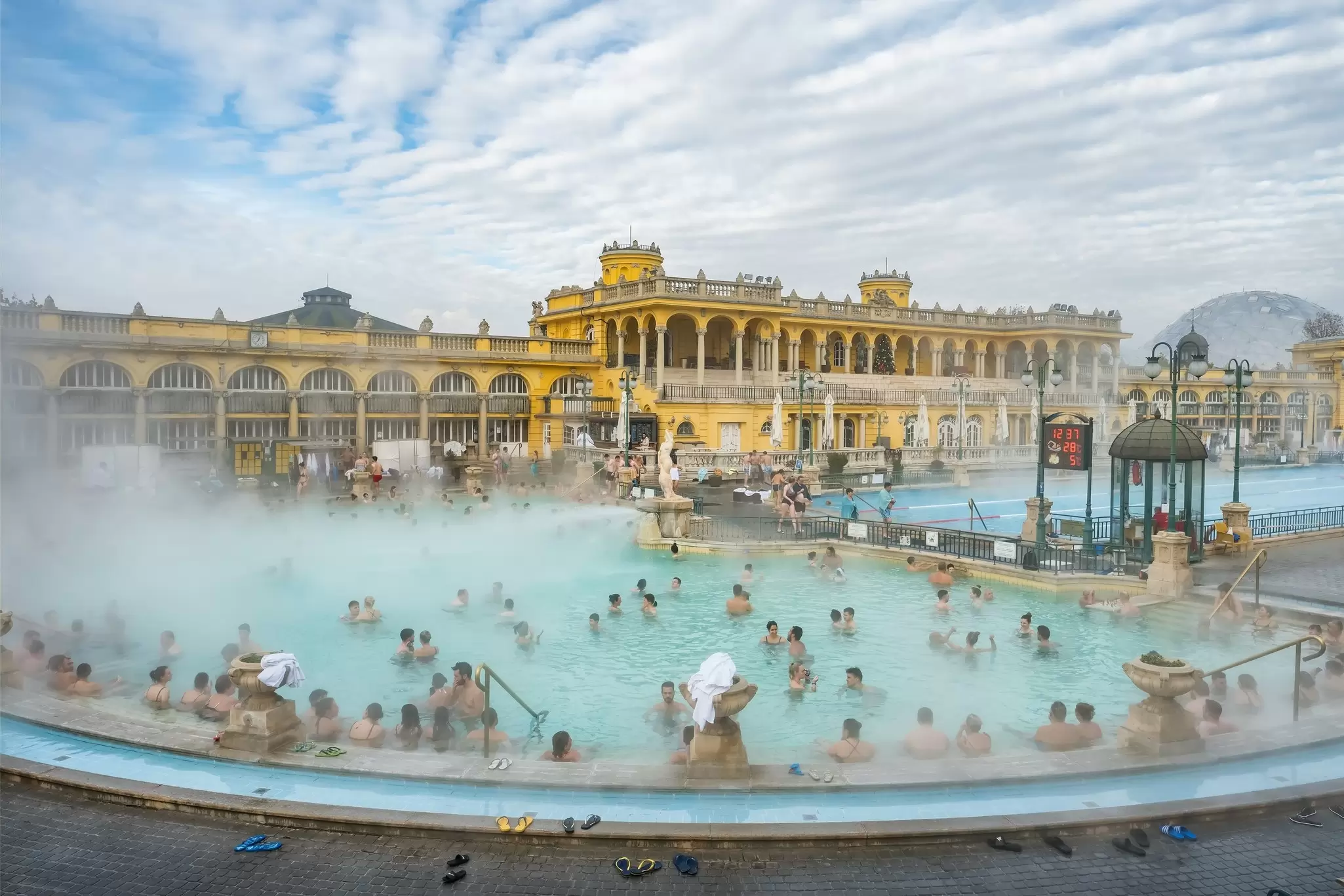 People in an outdoor pool with steam rising from it; a yellow building is in the background.