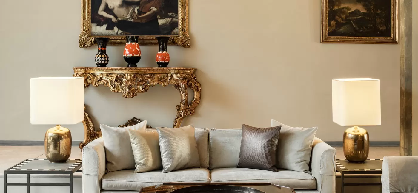 A hotel lobby sitting area with a large plush gray couch, side tables with gold lamps, Renaissance-style art and gold frames