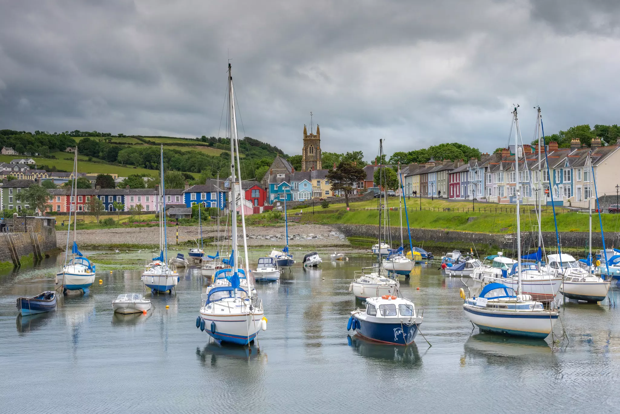 Small sailing and fishing boats moored in a harbor backed by a village of pastel-painted buildings and a church