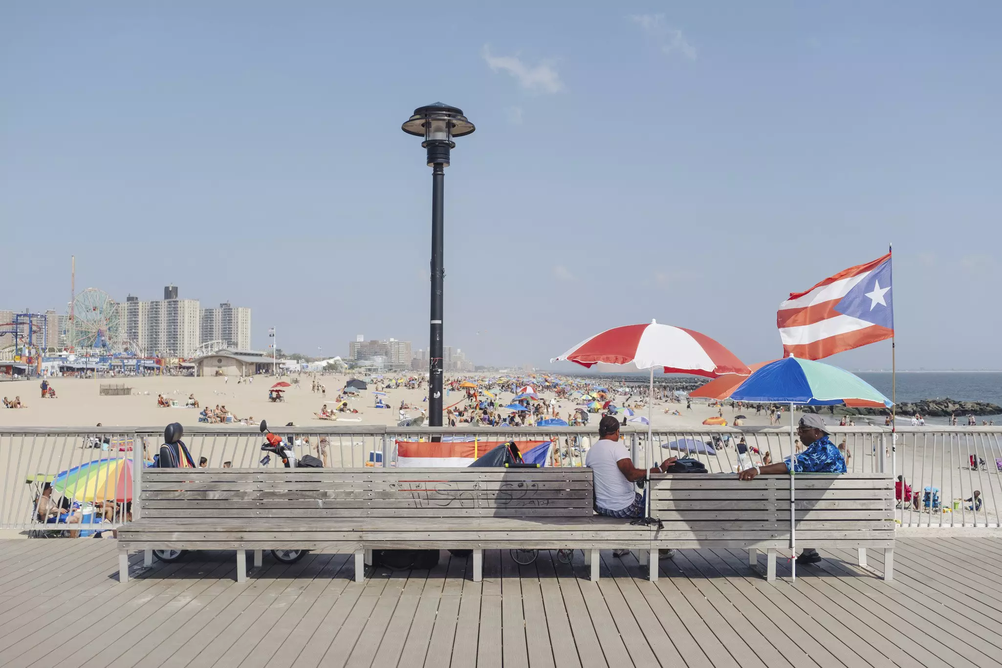 Two people sit on a bench under sun shades facing towards a wide sandy beach packed with sunbathers.