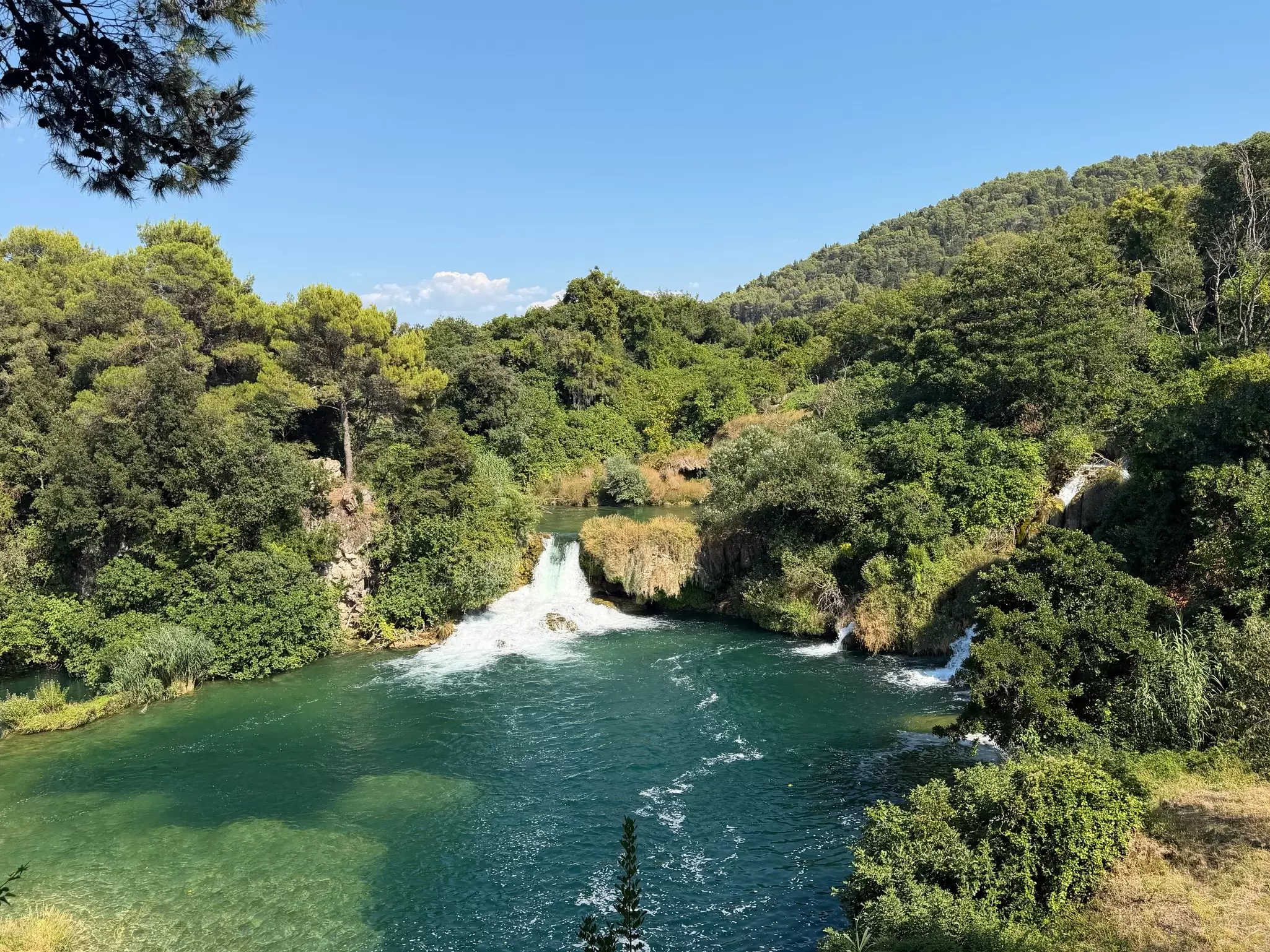 Large waterfall pools into a lake, it's surrounded by thick green trees. 