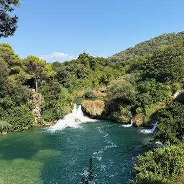 Large waterfall pools into a lake, it's surrounded by thick green trees. 