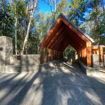 A timber-roofed entrance pavilion beside a stone wall at the start of a paved woodland trail.