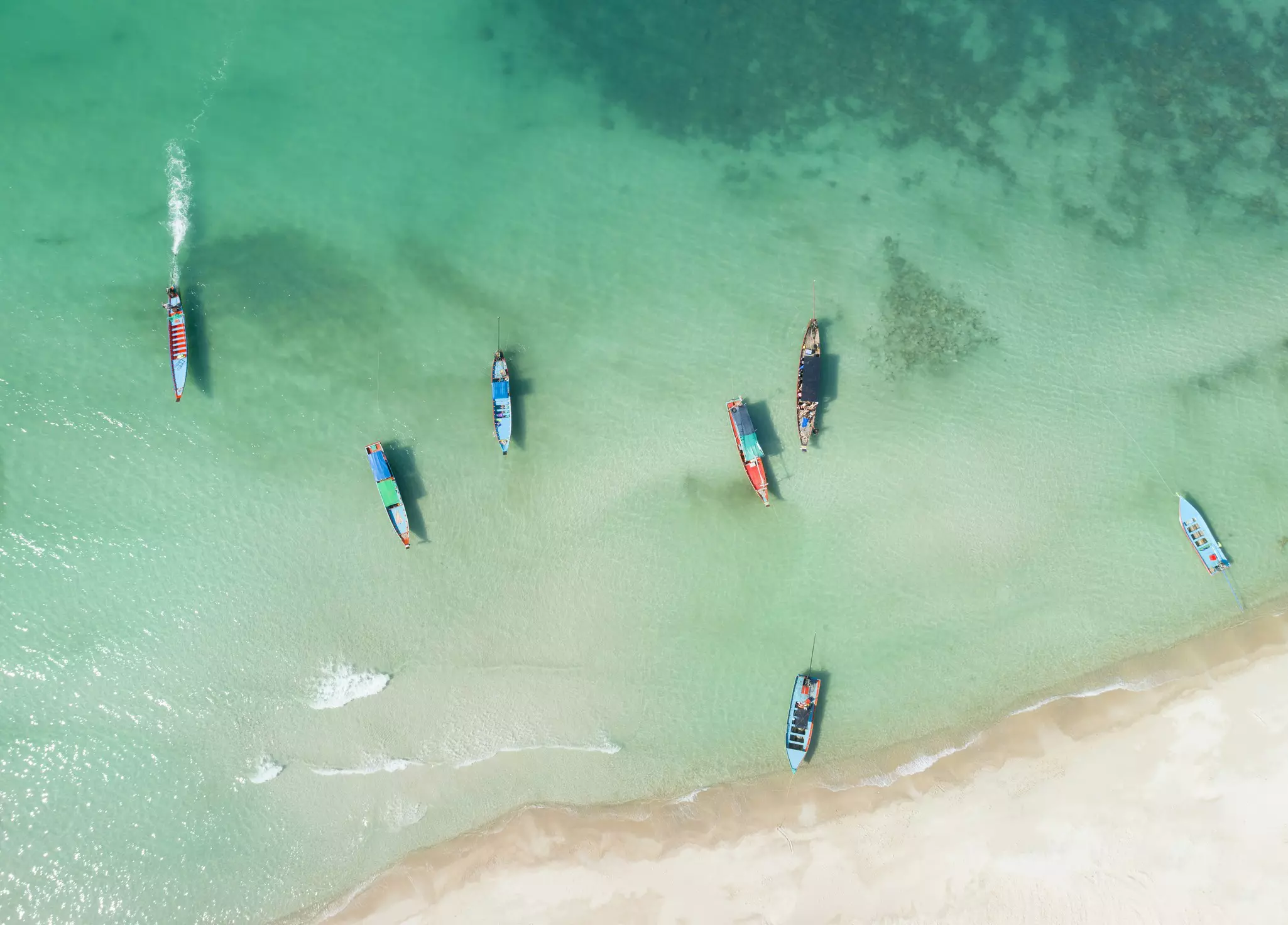 An aerial view of colorful fishing boats moored in shallow turquoise waters next to a white sand beach.