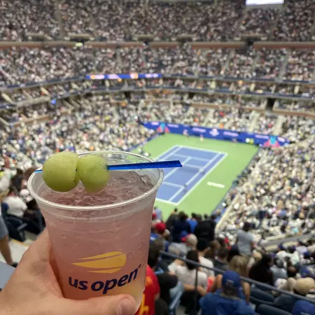 A hand holds up a cocktail with ice served in a plastic cup in front of a tennis court in a crowded arena at the US Open