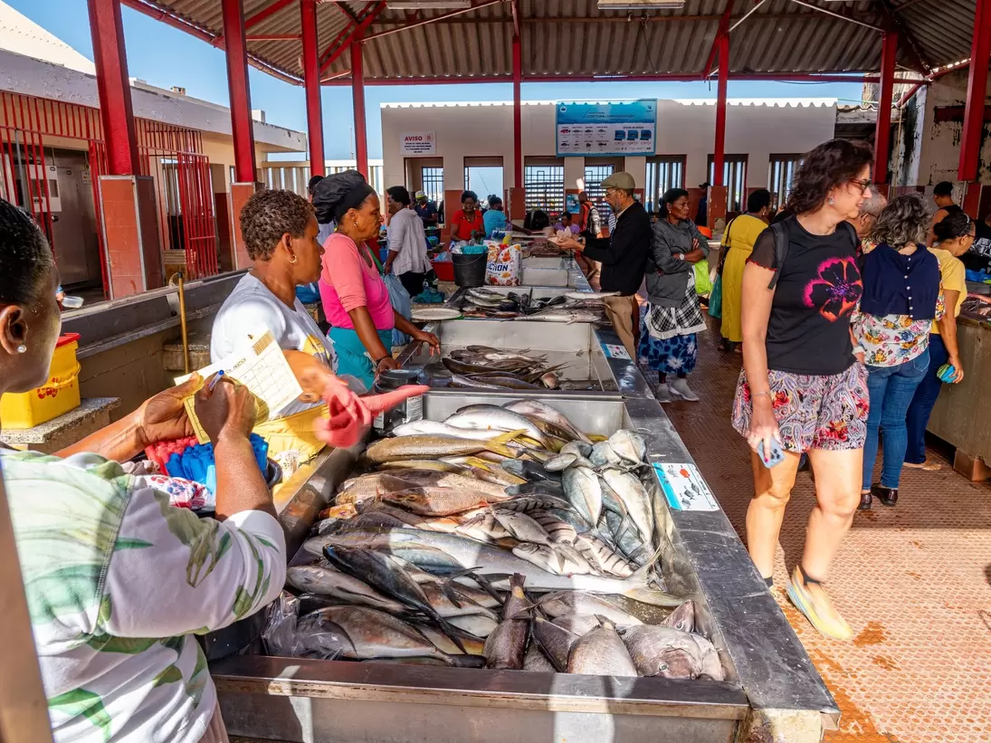 Covered market hall with red pillars, steel counters piled with fresh fish, vendors and shoppers walking along a tiled aisle