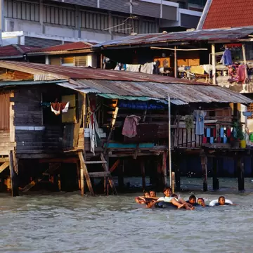Children floating on rubber rings in a brown river beside weathered wooden houses on stilts with laundry hanging out.