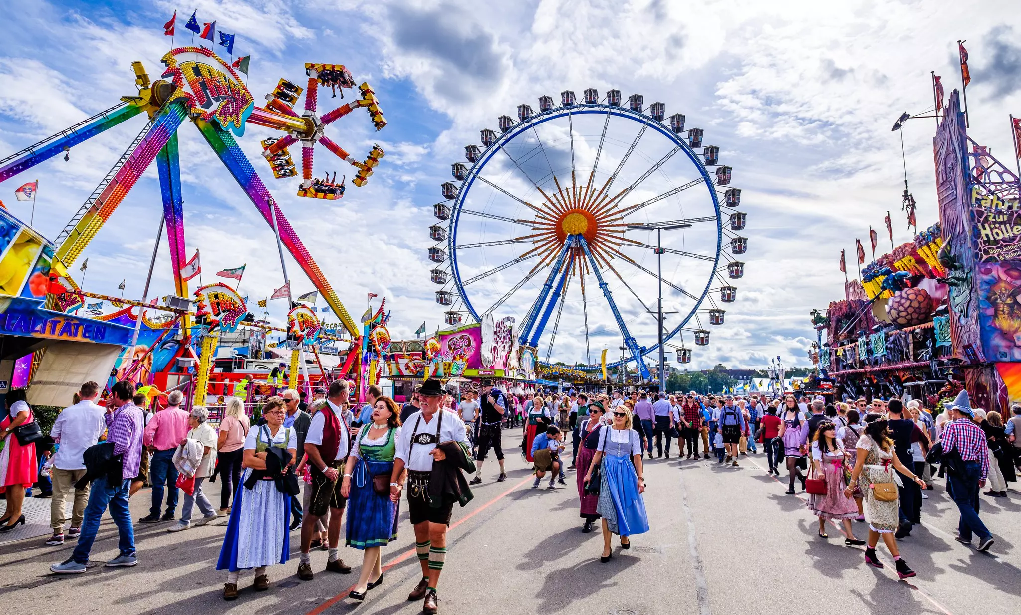 People walking the fairway in front of a Ferris wheel and other rides at a fairground