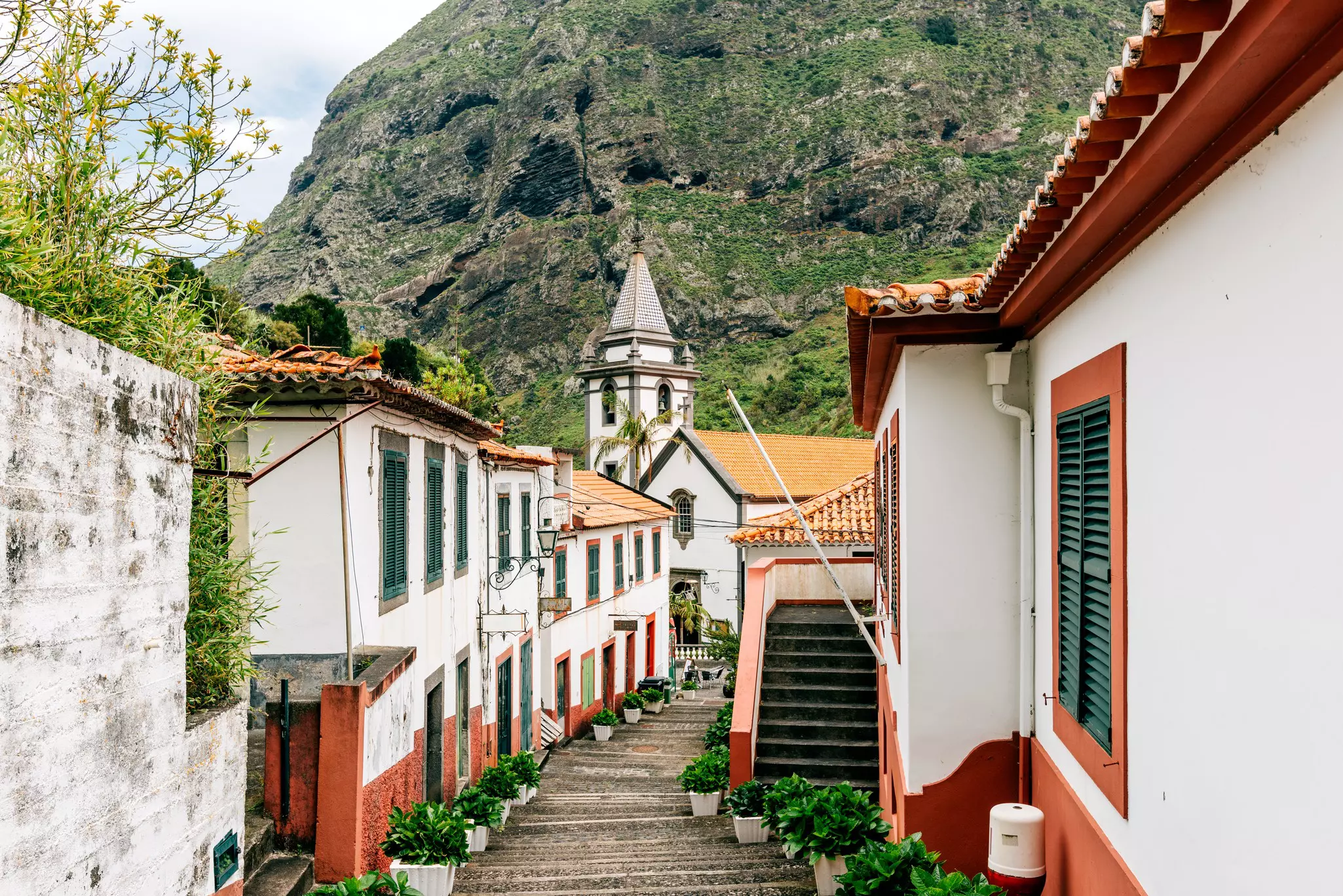 Stepped stone walkway through a mountain town with white buildings and red trim leads toward a church.