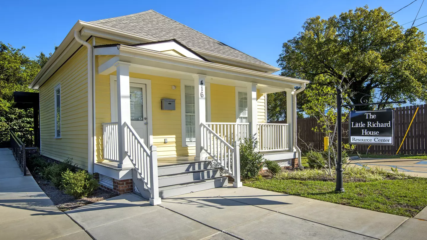 A small wooden house with a veranda painted yellow. A sign hanging outside says The Little Richard House