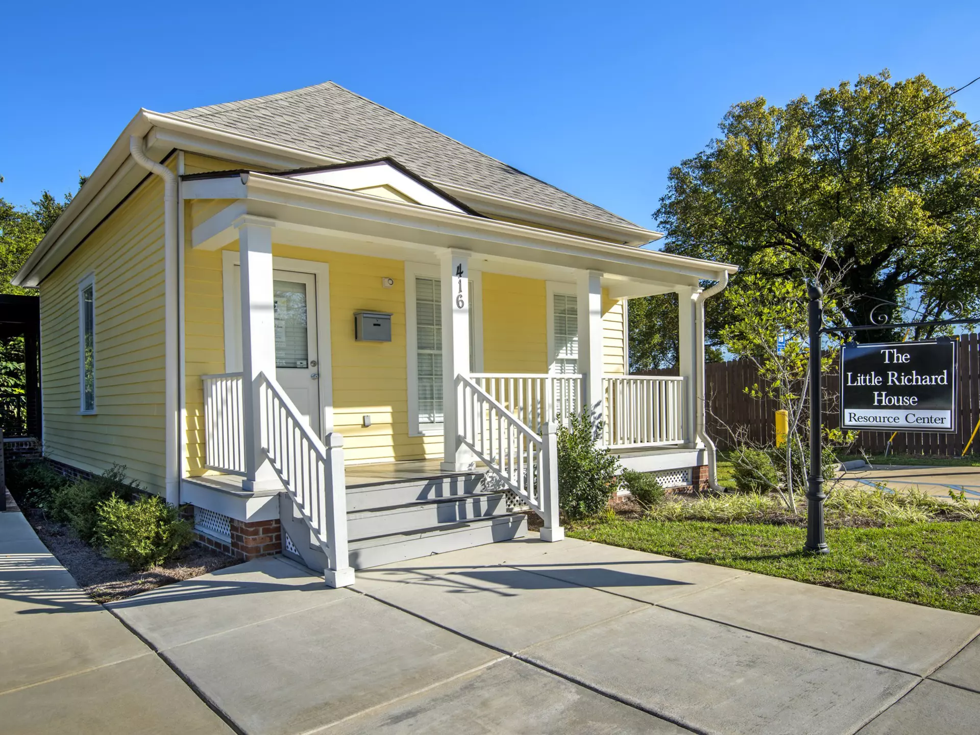A small wooden house with a veranda painted yellow. A sign hanging outside says The Little Richard House