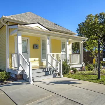 A small wooden house with a veranda painted yellow. A sign hanging outside says The Little Richard House