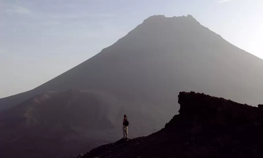 Hiker on a dark lava ridge facing the hazy volcanic cone of Pico do Fogo on Fogo island, Cabo Verde