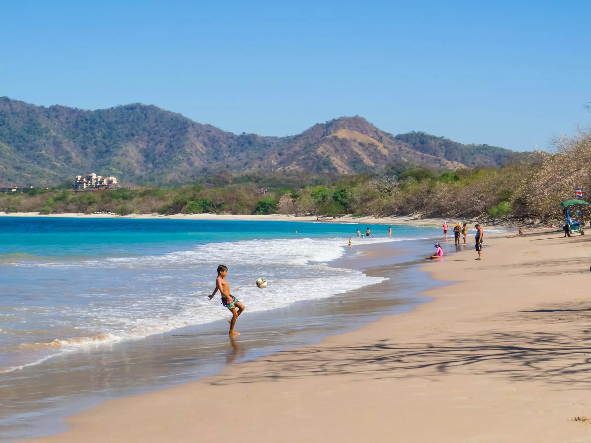 A teenager kicks a soccer ball on a wide tropical beach.