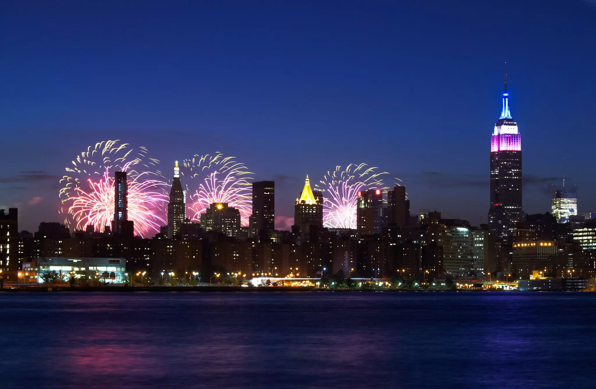 New York City skyline with the Empire State Building -- topped in red, white and blue -- at night with fireworks bursting behind them.