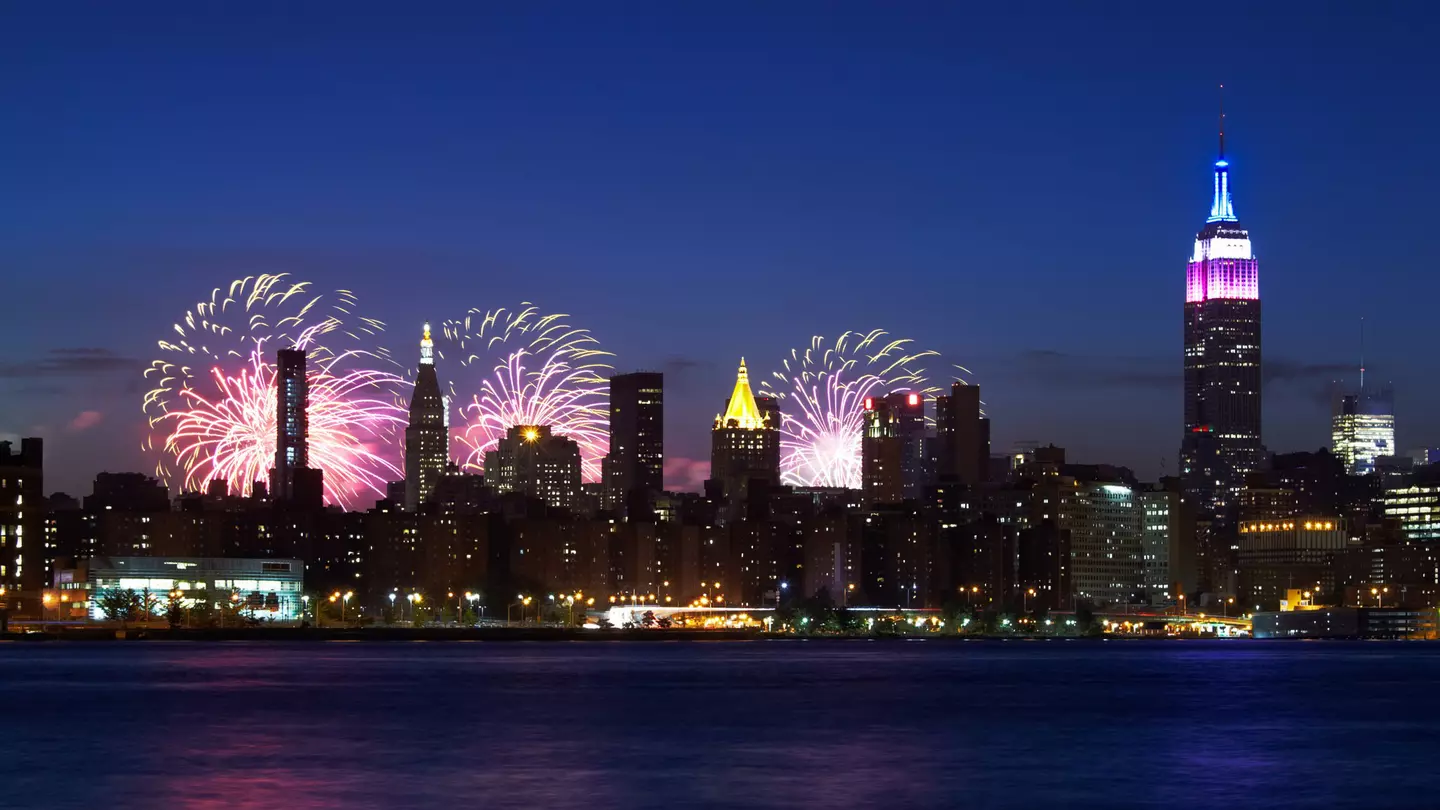 New York City skyline with the Empire State Building -- topped in red, white and blue -- at night with fireworks bursting behind them.