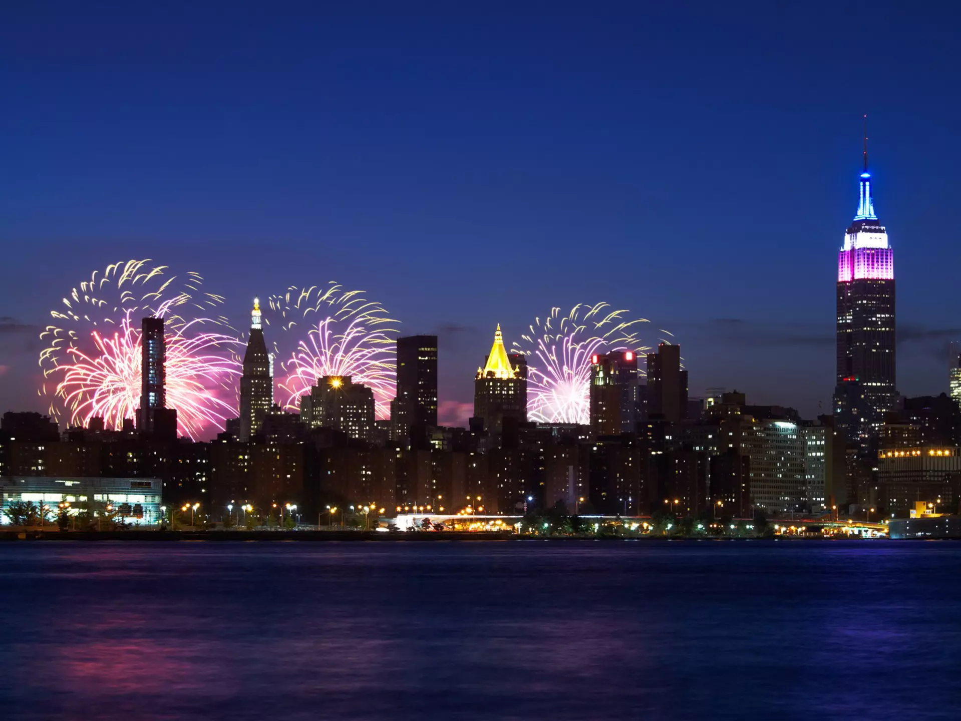 New York City skyline with the Empire State Building -- topped in red, white and blue -- at night with fireworks bursting behind them.