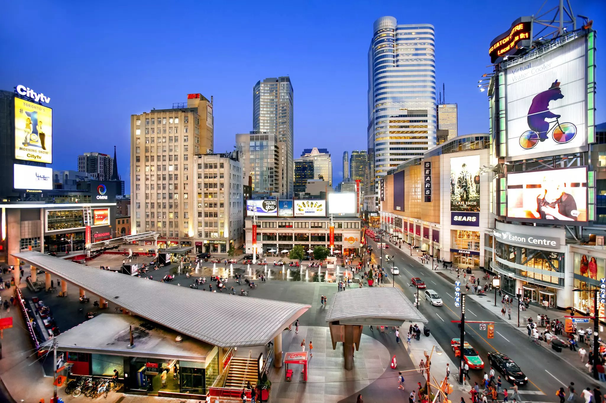 A brightly lit city square with TV screens showing advertising and people sat near pavement fountains
