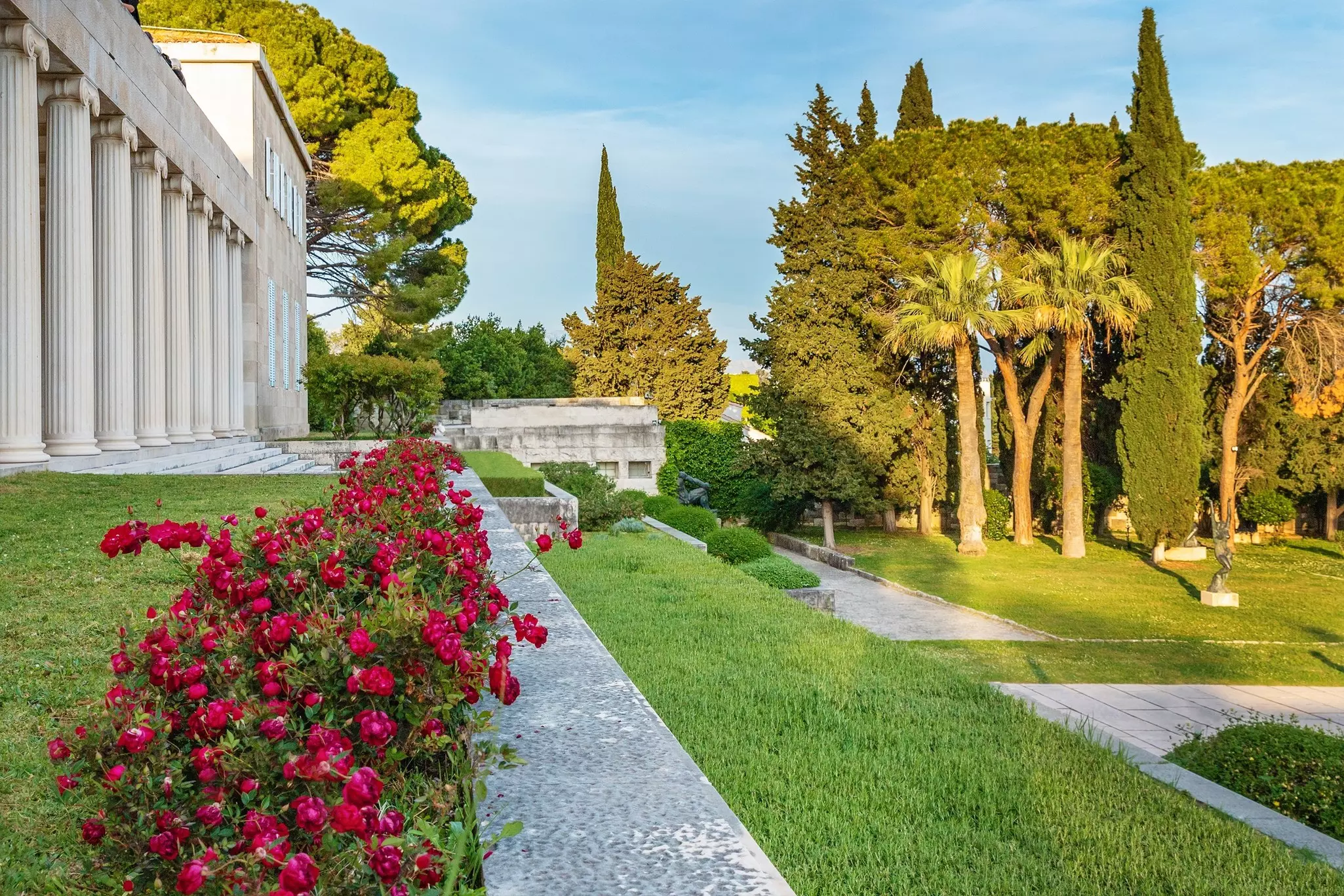 Beautiful landscaped gardens with sculptures outside a vast villa on a sunny day.
