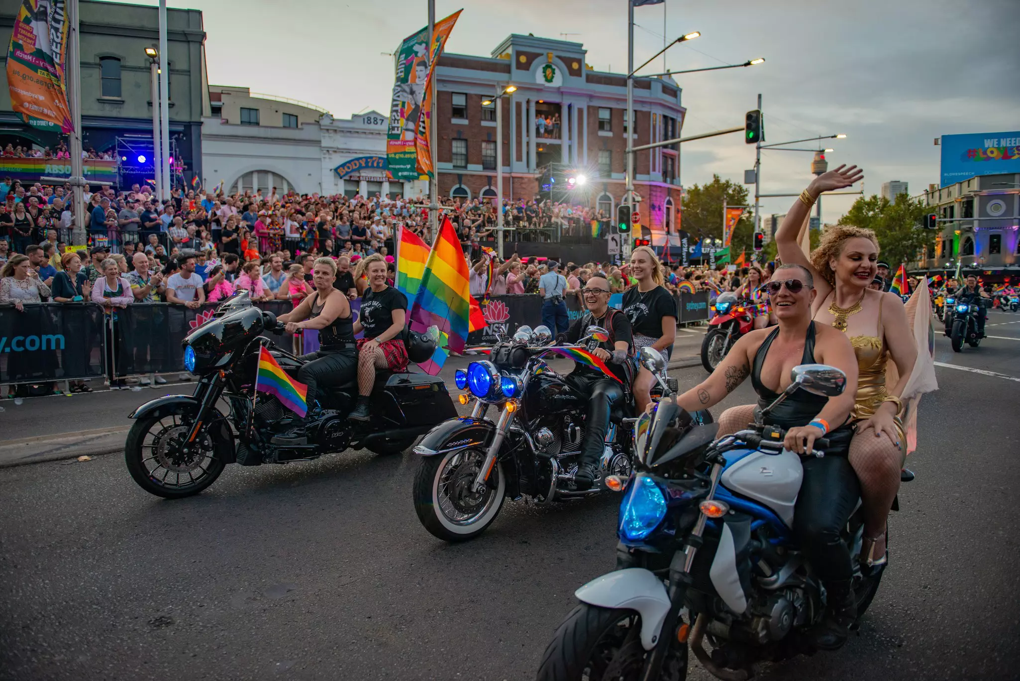 People on motorcycles with rainbow flags on the back wave at spectators on a parade route