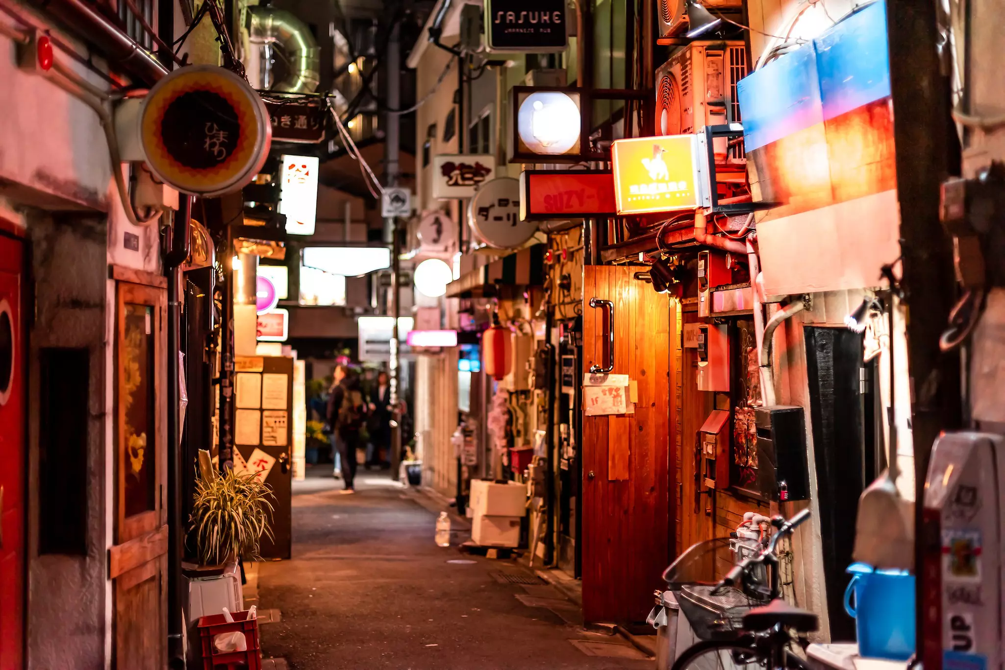 An alley in a city is lined with bars with illuminated signs.