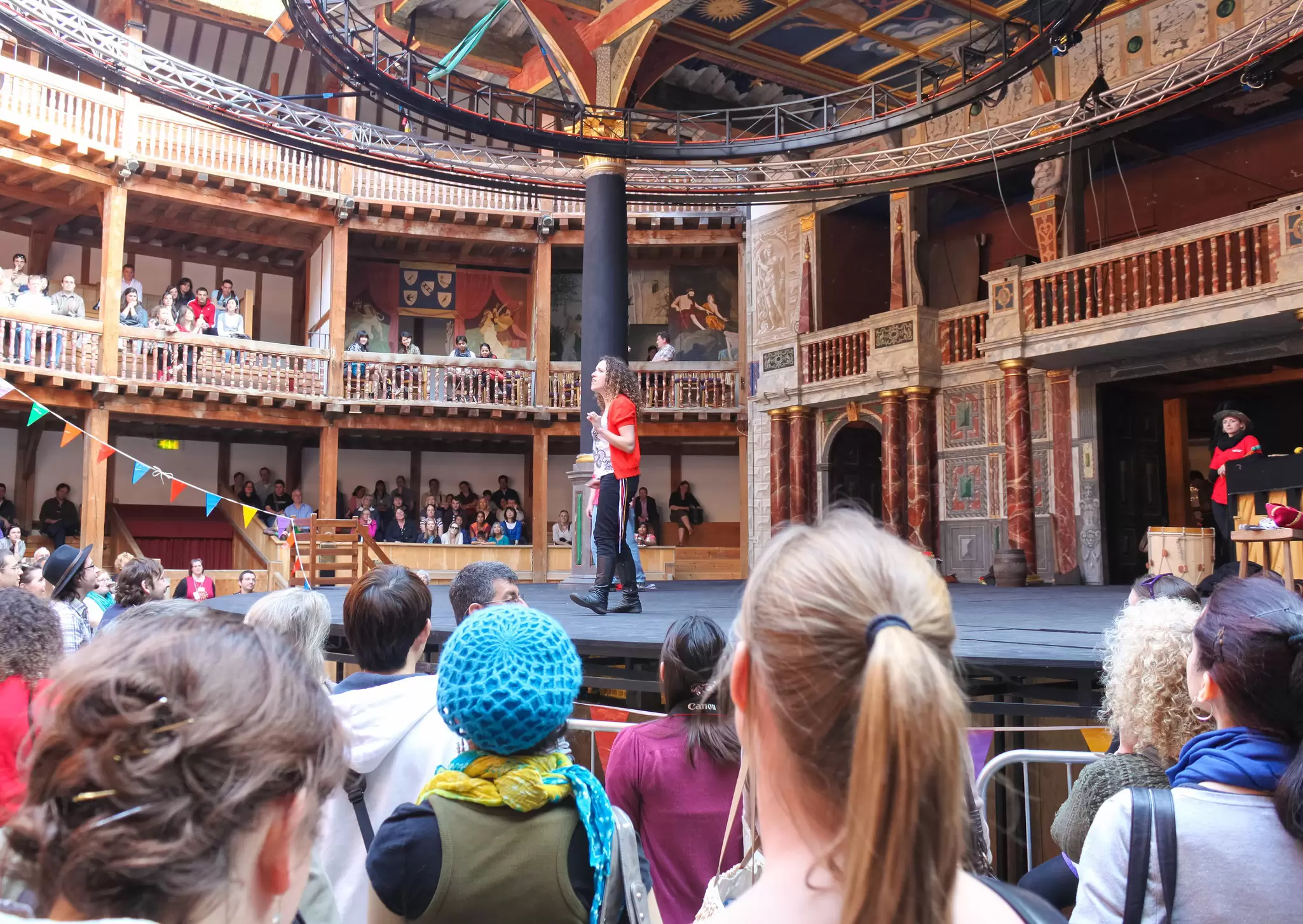 A crowd of people standing below a stage in a wooden theater. A person on the stage addresses the crowd