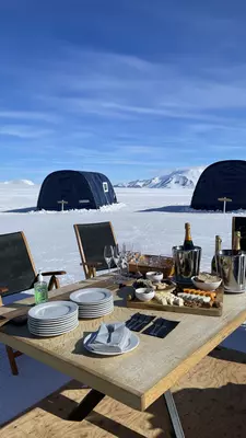 A table in the snow on a sunny day laden with Champagne bottles and sharing plates of food.