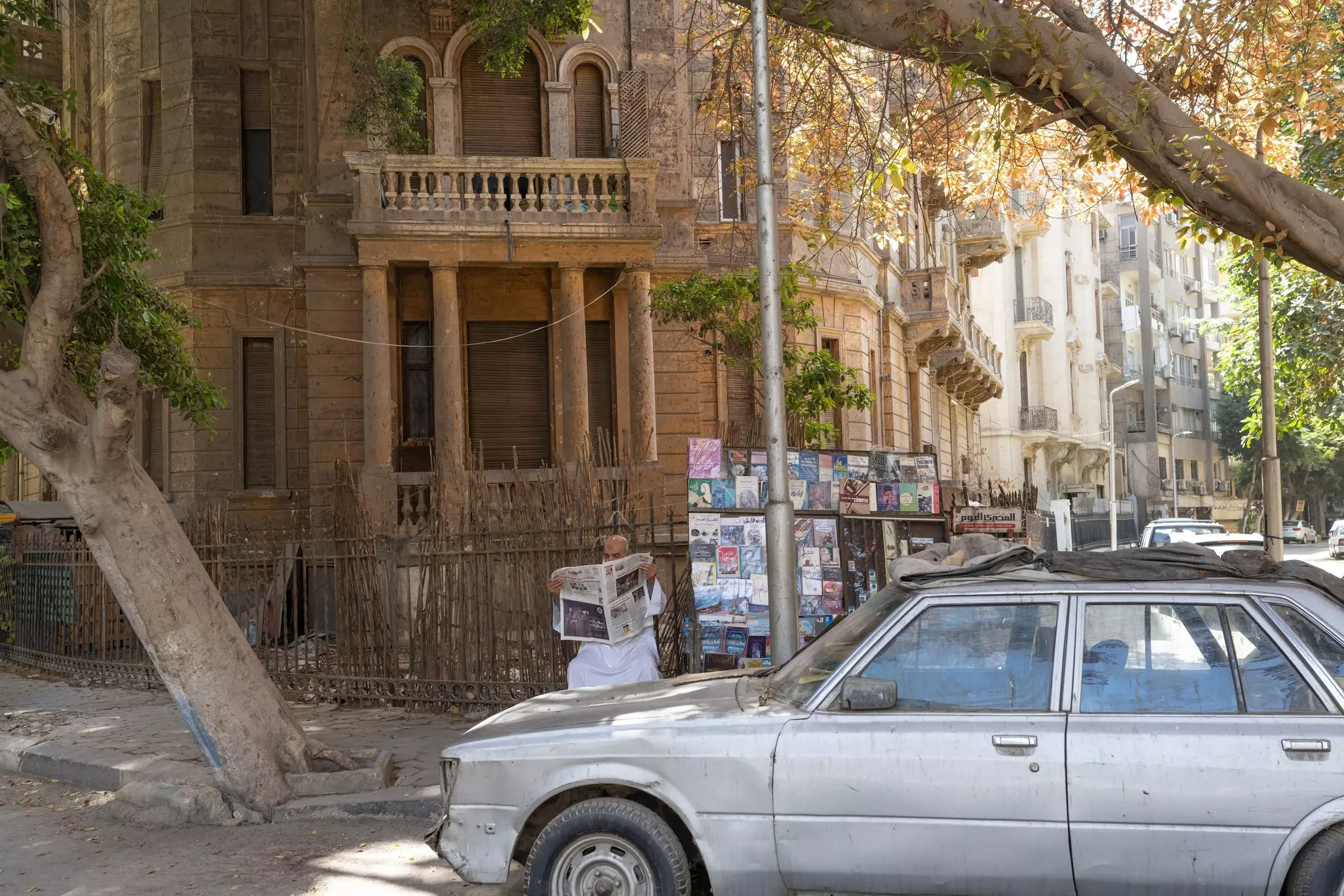 A man partially obscured by a parked gray car is seated while reading a newspaper on a shaded street corner beside a stall selling books and magazines in the Cairo neighborhood of Garden City.