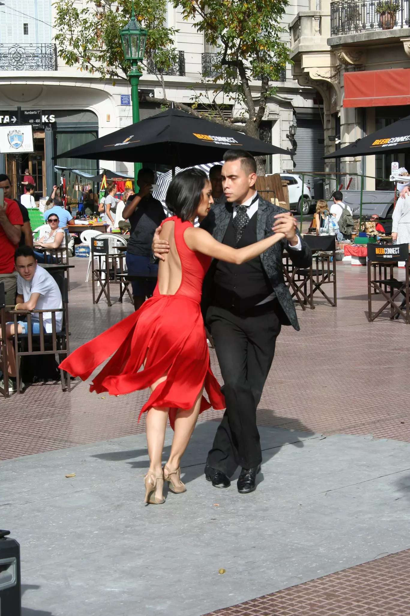 A woman in a red dress and a man in a black three-piece suit dance the tango on a plaza as people watch