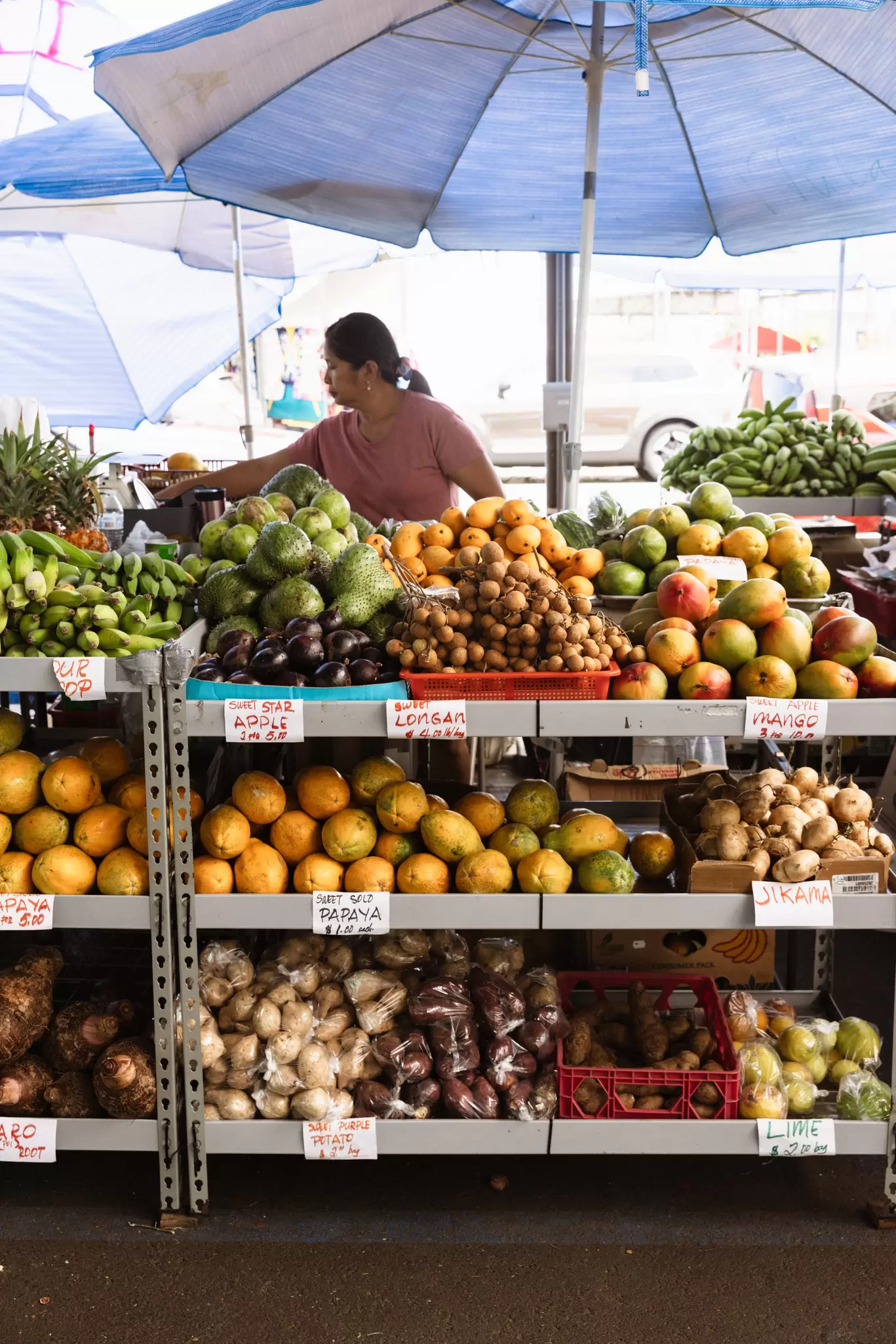 A woman under a sunshade at a market stall filled with fresh fruits and vegetables in Hilo, Hawaiʻi.