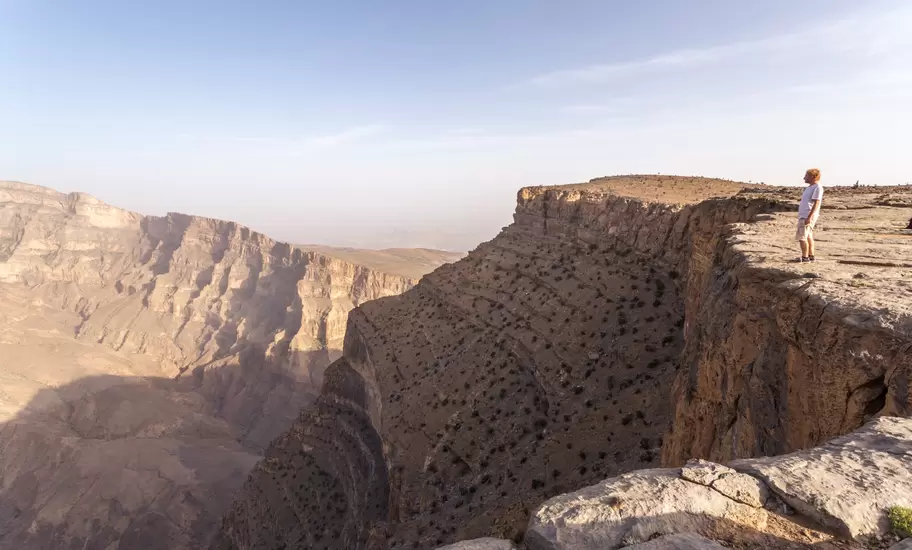 Man in white T-shirt standing on a limestone cliff edge above a deep layered canyon in Oman's Hajar Mountains at golden hour