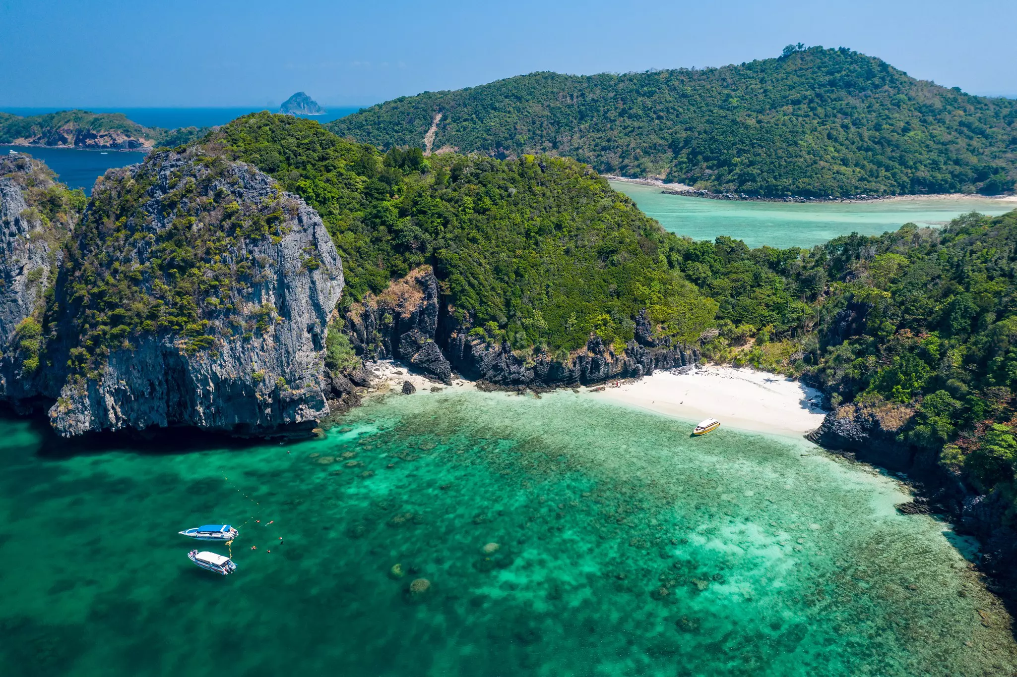 An aerial view of a cove with a small beach, hills and a rock formation covered in lush trees. Two boats are moored in the shallow water off the beach.