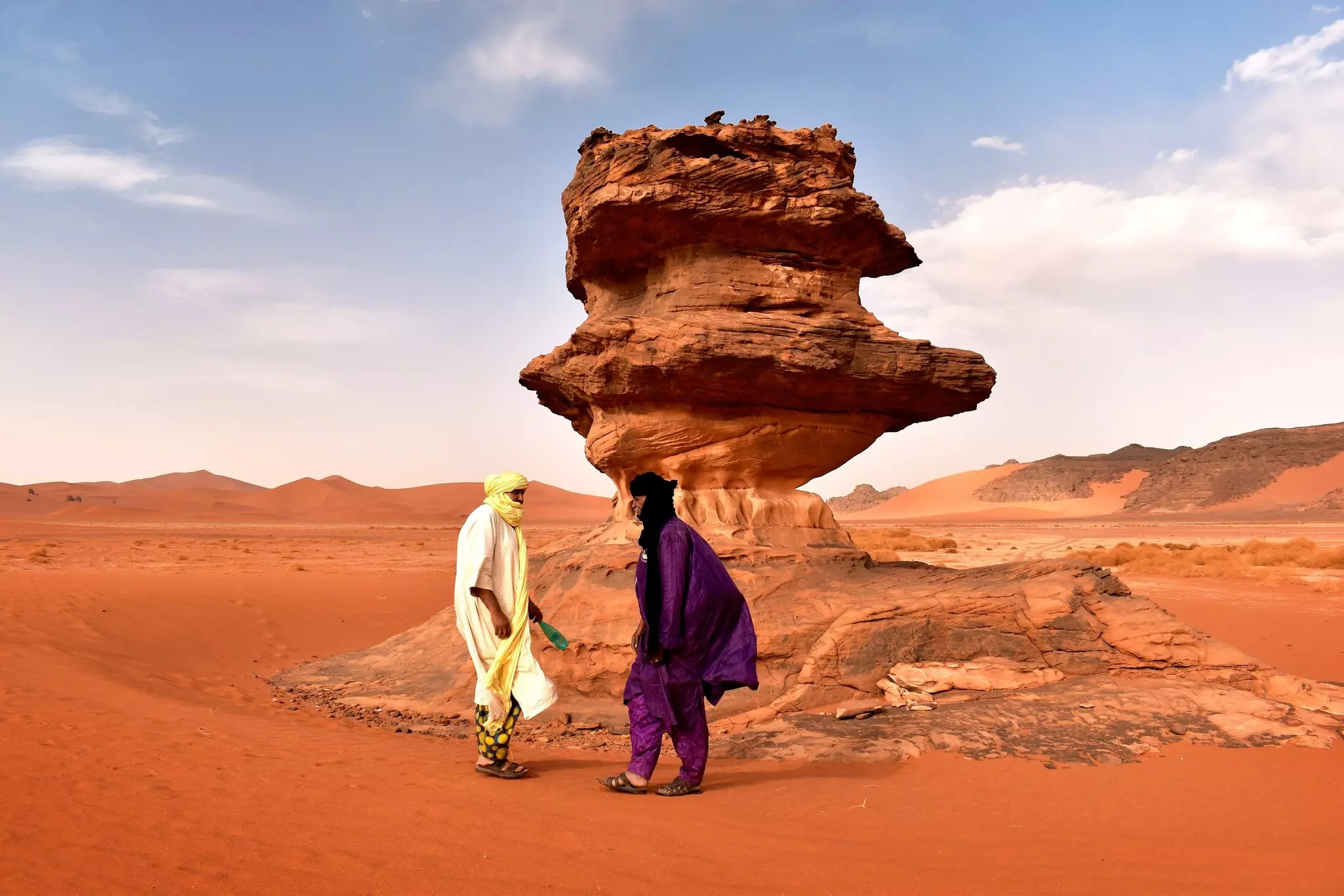 Two Tuareg guides stand by a rock formation in the middle of a red sand plateau in Algeria.