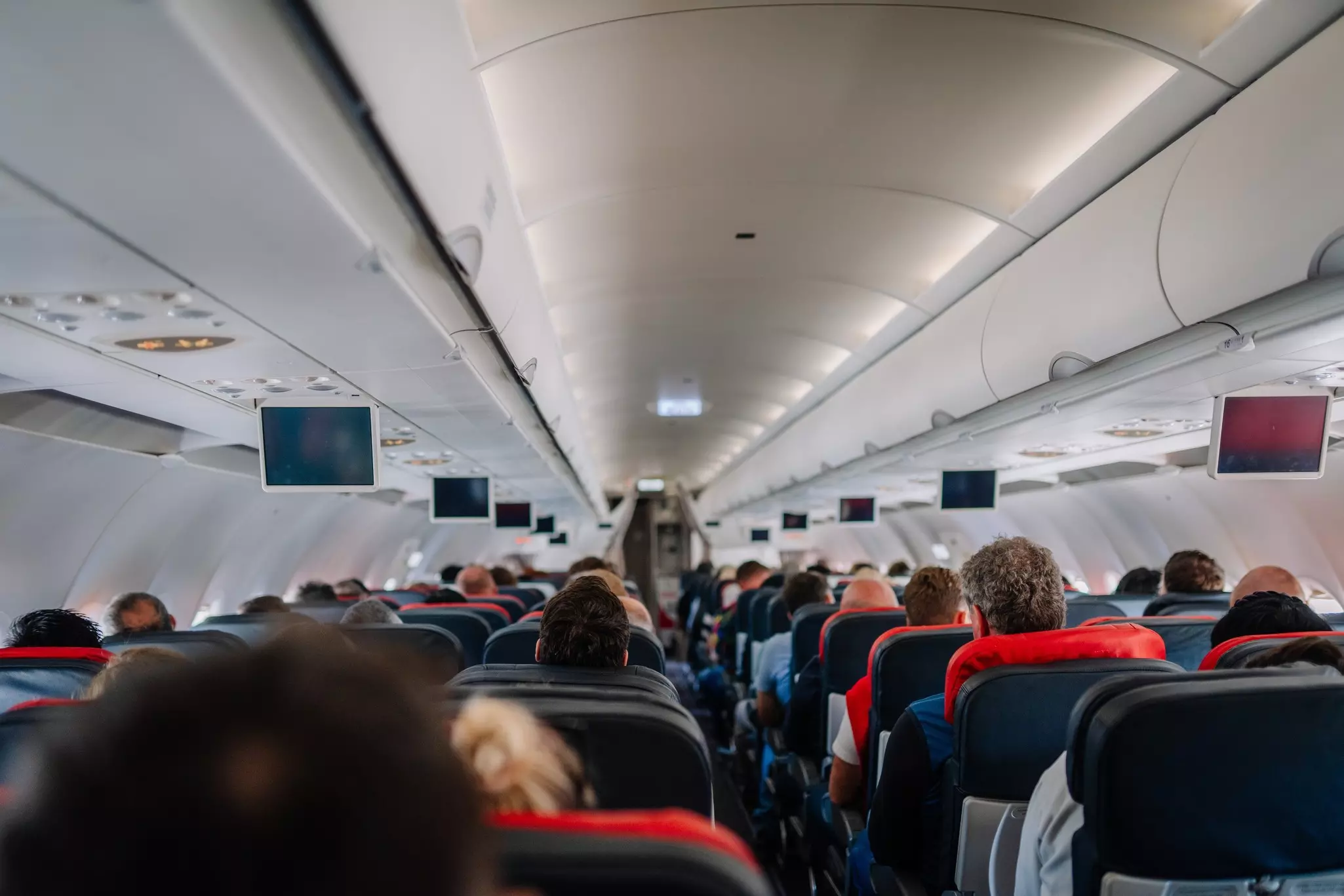Interior view of an airplane cabin filled with passengers seated during a flight. Overhead compartments, screens, and lighting are visible