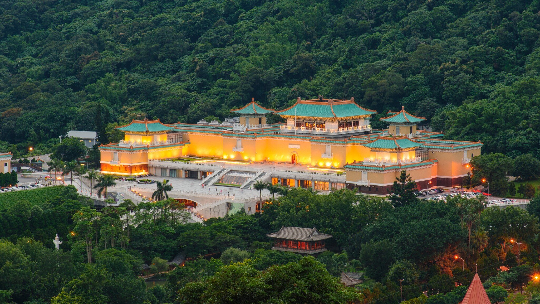 The National Palace Museum lit up at dusk against a backdrop of dark green forest in Taipei, Taiwan.