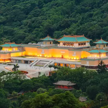 The National Palace Museum lit up at dusk against a backdrop of dark green forest in Taipei, Taiwan.