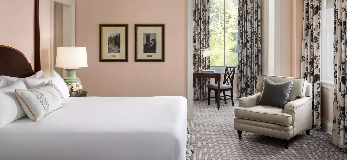 A hotel bedroom with a king-size bed, floral curtains, a chair, desk, and equestrian artwork