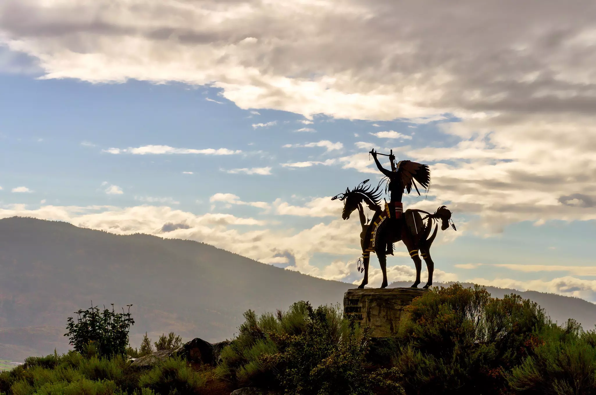 A silhouetted statue of an Indigenous warrior with moutains and clouds in the distance.