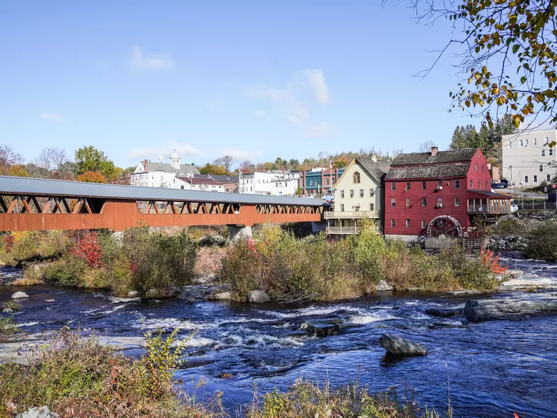 A covered bridge over a river with small areas of rapids leads to a New Hampshire town with simple historic-looking buildings.