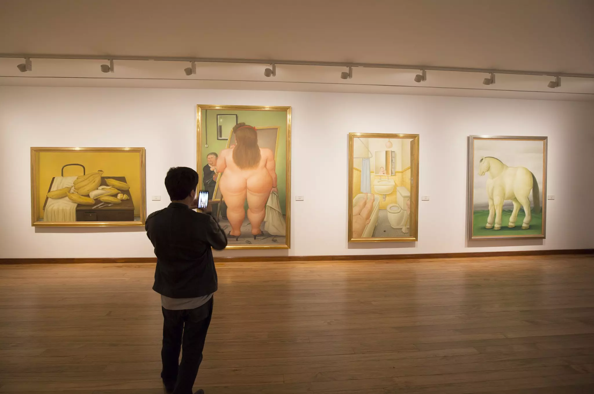 A man is seen from behind taking a picture of four paintings hanging on the wall of a museum gallery.